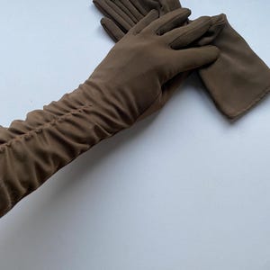 May include: A pair of brown gloves. One glove is laid flat, while the other is partially covering it. The gloves are made of a soft material and have a gathered detail on the arm.