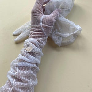 May include: A pair of white sheer opera-length gloves featuring a delicate diamond-shaped mesh pattern. The fabric is lightweight and semi-transparent, extending past the wrist with a gathered, ruched texture along the forearm and fingers.