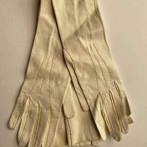 May include: A pair of cream-colored leather gloves. The gloves are long, extending past the wrist, and feature detailed stitching along the fingers and seams. The leather has a slightly textured appearance, and the gloves are laid flat.
