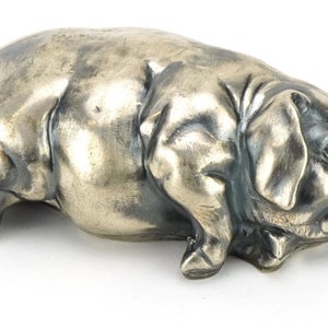 Puede incluir: Una figurita de cerdo de plata con ojos de vidrio rojo. El cerdo está acostado sobre su vientre con la cabeza girada hacia un lado.