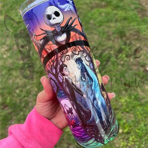 Puede incluir: Un vaso de acero inoxidable con un dise&ntilde;o colorido que presenta personajes de "Pesadilla antes de Navidad" y "La Novia Cad&aacute;ver". El dise&ntilde;o incluye a Jack Skellington, Emily y Victor, sobre un fondo espeluznante. El vaso es sostenido por una persona.