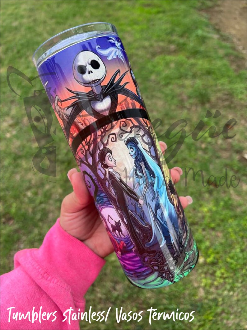 Puede incluir: Un vaso de acero inoxidable con un dise&ntilde;o colorido que presenta personajes de "Pesadilla antes de Navidad" y "La Novia Cad&aacute;ver". El dise&ntilde;o incluye a Jack Skellington, Emily y Victor, sobre un fondo espeluznante. El vaso es sostenido por una persona.