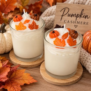May include: Two glass jar candles filled with off-white wax, topped with white whipped wax, orange and brown pumpkin-shaped wax embeds, and orange maple leaf wax accents. A wooden sign behind the candles displays the text Pumpkin CASHMERE.