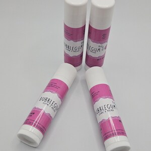 May include: Four white lip balm tubes with pink labels. The labels read "Bubblegum Comme & Macher". The tubes are arranged in a circular pattern against a white background. The lip balms are a cosmetic product.