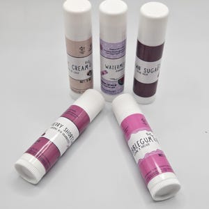 May include: Five lip balm tubes with white caps and labels. The labels feature text like "Cream de Coco", "Watermelon", "Brown Sugar", "Berry Shortcake", and "Bubblegum". The tubes are arranged on a white surface.