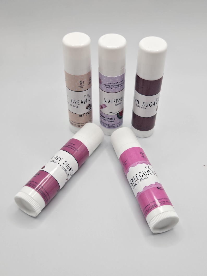 May include: Five lip balm tubes with white caps and labels. The labels feature text like "Cream de Coco", "Watermelon", "Brown Sugar", "Berry Shortcake", and "Bubblegum". The tubes are arranged on a white surface.