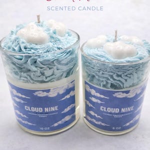 May include: Two glass jar candles with blue frosting and white cloud-shaped wax accents. The candles have a label that reads "Cloud Nine" with a blue sky and cloud design. One candle is 283g, the other is 227g.