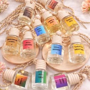 Op de afbeelding: Een verzameling kleine, heldere glazen flesjes met houten doppen en kleurrijke etiketten. De etiketten tonen teksten als "Apple Orchard" en "Snowy Morning". Elk flesje hangt aan een goudkleurig koord, waarschijnlijk luchtverfrissers.