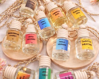 May include: A collection of small, clear glass bottles with wooden caps and colorful labels. The labels feature text like "Pumpkin Spice," "Apple Orchard," and "Snowy Morning." Each bottle is suspended by a gold-colored cord, suggesting they are air fresheners.