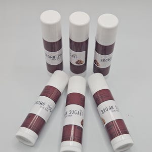 May include: Six lip balm tubes with white caps and brown and white labels. The labels read "BROWN SUGAR SUCRE ROUX" and feature a small graphic of brown sugar. The tubes are arranged on a white surface.