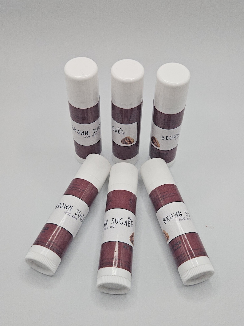 May include: Six lip balm tubes with white caps and brown and white labels. The labels read "BROWN SUGAR SUCRE ROUX" and feature a small graphic of brown sugar. The tubes are arranged on a white surface.