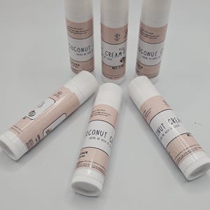 May include: A collection of six white lip balm tubes with pink labels. The labels read "COCONUT CREAM" and "BALM" in a stylized font. The tubes are arranged on a light surface.