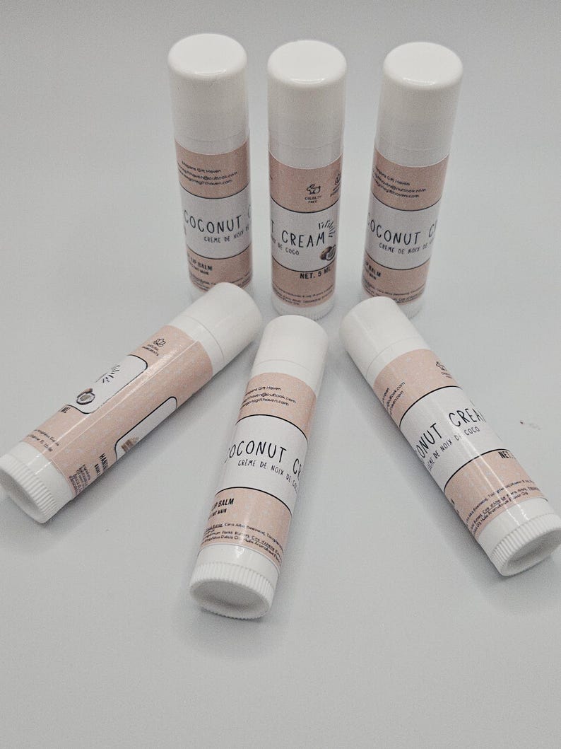 May include: A collection of six white lip balm tubes with pink labels. The labels read "COCONUT CREAM" and "BALM" in a stylized font. The tubes are arranged on a light surface.