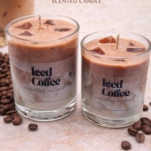 Peut inclure: Deux bougies en verre avec un aspect café, étiquetées "Iced Coffee Scented Candle". Les bougies ont une couche supérieure marron avec ce qui semble être des glaçons et une couche inférieure plus claire. Des grains de café sont éparpillés.