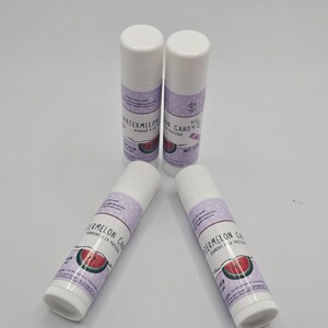 May include: Four white lip balm tubes with white caps are arranged on a white surface. Each tube has a purple label with the words "Watermelon Candy" and a watermelon slice graphic. The net weight is 0.15 oz.