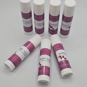 May include: Six lip balm tubes with white caps and bases, and a magenta band with text. The text includes "Raspberry Shortcake" and "Handmade Lip Balm". The tubes are arranged on a white surface.