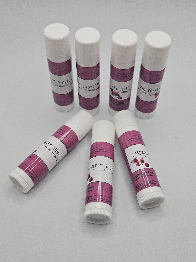 May include: Six lip balm tubes with white caps and bases, and a magenta band with text. The text includes "Raspberry Shortcake" and "Handmade Lip Balm". The tubes are arranged on a white surface.