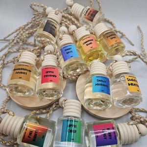 Puede incluir: Una colección de pequeños frascos de vidrio transparente con tapas de madera y etiquetas con varios nombres de fragancias, como "Pumpkin Spice" y "Cinnamon Buns". Cada frasco está unido a un cordón decorativo.