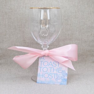 May include: A clear glass wine glass with a gold rim tied with a pink ribbon and a gift tag that reads "A Toast to the Host! Clink!"  Macy and Trevor Rouke.