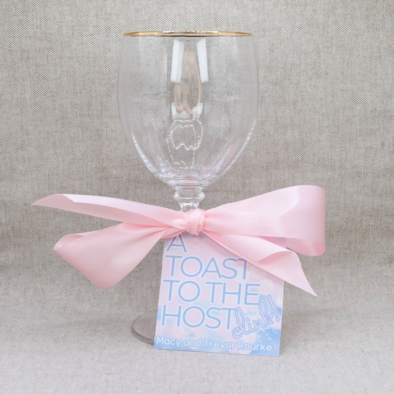 May include: A clear glass wine glass with a gold rim tied with a pink ribbon and a gift tag that reads "A Toast to the Host! Clink!"  Macy and Trevor Rouke.