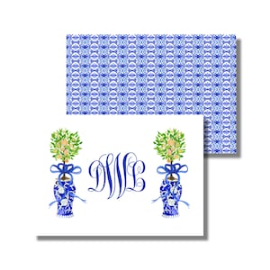 Puede incluir: Dos jarrones de porcelana azul y blanco con una cinta azul y un pequeño árbol verde con flores amarillas en su interior. Los jarrones están sobre un fondo blanco con el monograma "DWL" en azul.