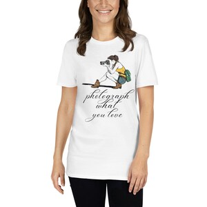 以下が含まれることがあります： カメラで写真を撮っている女性のグラフィックがプリントされた白いTシャツ。グラフィックの下には「愛するものを写真に撮る」というテキストがプリントされています。