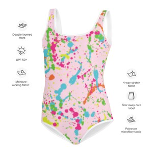 May include: A pink one-piece swimsuit with a colorful paint splatter design. The swimsuit has a white trim around the neckline and armholes. It is made of polyester microfiber fabric and has a tear-away care label.