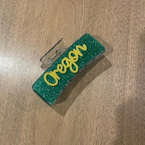 May include: A clear plastic hair claw clip with a rectangular, glittery green base. The word "Oregon" is written in yellow script across the top. The clip is laying on a light brown wooden surface.