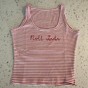 May include: A red and white striped tank top with the embroidered text "Roll Tide" in red script. The tank top has a ribbed texture and a scoop neckline.