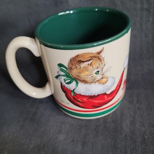 Puede incluir: Taza de cerámica color crema con interior y asa verdes. La taza presenta una ilustración festiva de un gatito con un gorro de Papá Noel. El gorro es rojo con ribete blanco y el gatito tiene ojos verdes. Un lazo verde está atado alrededor del cuello del gatito.