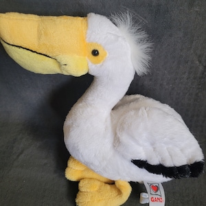 Puede incluir: Pelícano de peluche blanco con pico y patas amarillas. El pelícano tiene una franja negra en la espalda y un pequeño mechón de pelo blanco en la cabeza. El peluche tiene una etiqueta roja en forma de corazón con la palabra "Ganz" en ella.