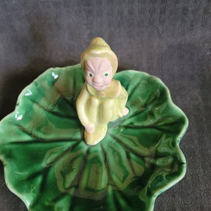 Può includere: Un piatto in ceramica verde a forma di foglia con una piccola statuina di un folletto verde seduto sopra.