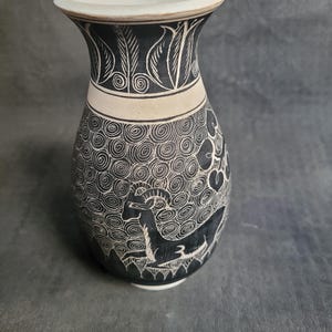 Puede incluir: Un jarrón de cerámica blanca con diseños grabados en blanco y negro. El jarrón presenta un animal estilizado con cuernos y un patrón de espiral.