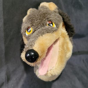 May include: A plush dog puppet with a black nose, yellow and black eyes, and a pink tongue. The puppet has brown and tan fur and is designed to fit over a hand for play.