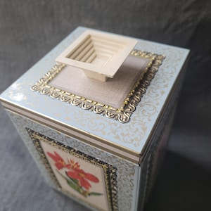 Könnte beinhalten: Eine quadratische, hellblaue Zierbox mit floralem Design und goldfarbenem Rand. Die Box zeigt ein gerahmtes Bild einer roten Blume und ein mehrstufiges, cremefarbenes Quadrat oben. Die Box ist wahrscheinlich aus Metall.