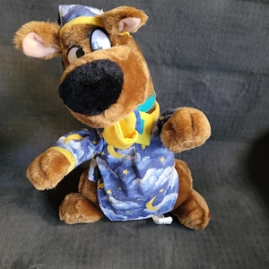 Puede incluir: Un Scooby Doo de peluche marrón que lleva una bata de dormir azul y amarilla con estrellas y lunas. Lleva una gorra de dormir azul y amarilla.