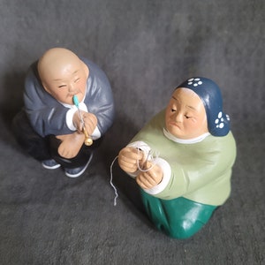 Puede incluir: Dos figuritas de cerámica de personas mayores. El hombre lleva una túnica gris y sostiene una pipa. La mujer lleva una túnica verde con un pañuelo de cabeza estampado en azul y blanco y sostiene un trozo de cuerda.