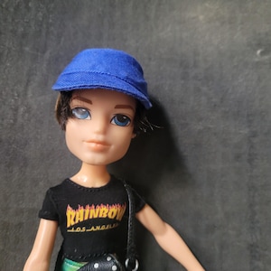 Könnte beinhalten: Eine blaue und schwarze Puppe mit blauen Augen, die eine blaue Baseballkappe, ein schwarzes T-Shirt mit dem Aufdruck "Rainbow Los Angeles" und eine schwarz-weiß gepunktete Tasche trägt.