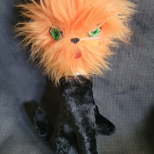May include: A handmade cat doll with a fluffy orange head, green eyes, and a black body. The cat's face has a small red tongue sticking out. The doll's body is black and has a hand-like shape.
