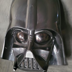 Puede incluir: Una máscara de Darth Vader de plástico negro con un diseño detallado. La máscara tiene una visera negra y una rejilla de boca negra.