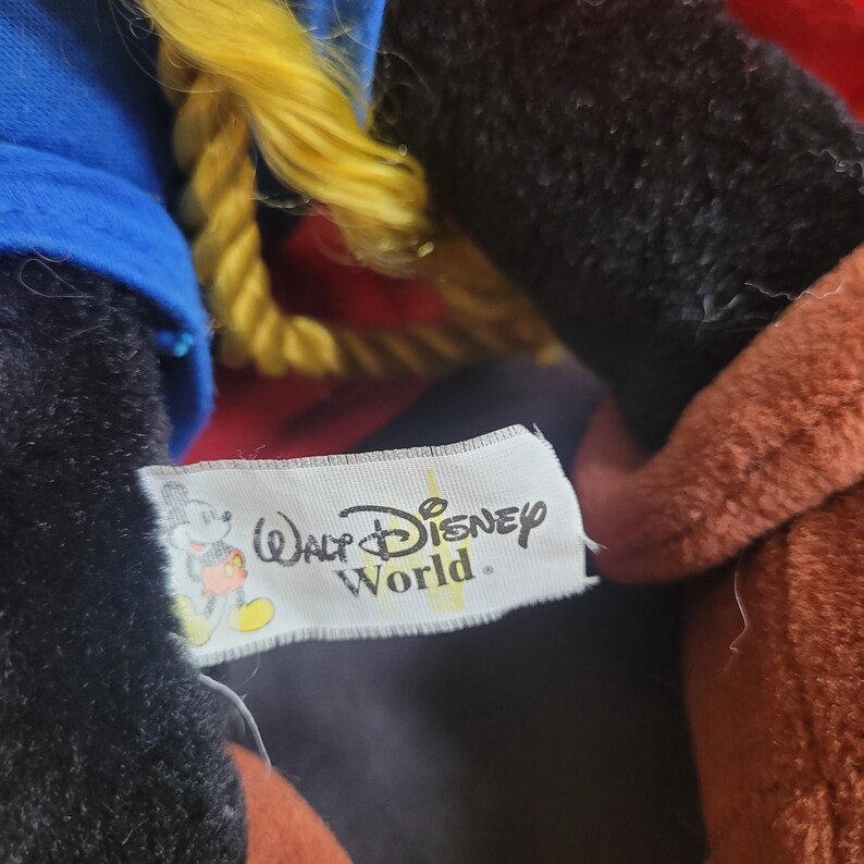 May include: Close-up of a plush toy with a blue hat, black fur, and a red and yellow outfit. A white tag with the "Walt Disney World" logo is visible.