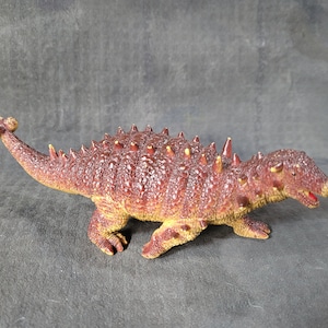 Może przedstawiać: Zabawka dinozaur w kolorze czerwonym i żółtym z kolcami na grzbiecie. Dinozaur stoi na szarym tle.