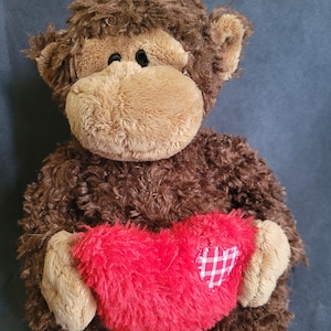 May include: A plush monkey toy with brown fur and a tan face and ears. The monkey is holding a red heart with a white plaid patch. The toy has black eyes and a friendly expression.