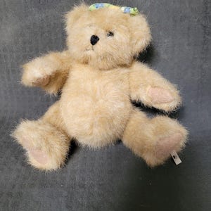 Puede incluir: Un oso de peluche marrón claro con una diadema floral azul y verde. El oso está sentado sobre una superficie gris.