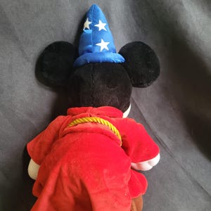 May include: A plush Mickey Mouse toy dressed as a sorcerer, viewed from the back. Mickey wears a red robe with a gold rope belt and a blue wizard hat with white stars. The toy has black ears and a black head.