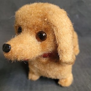 Puede incluir: Un perro de peluche marrón con ojos negros y un collar rojo. El perro está de pie sobre una superficie gris.