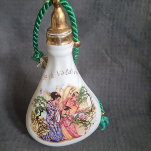 May include: White ceramic bottle with a green rope handle and a gold lid. The bottle features a painted scene of two people in traditional clothing standing in a garden with trees and flowers. The text "Valdivia" is written on the bottle.