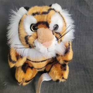 May include: A plush tiger toy with orange and black stripes, white fur around its face, and black whiskers. The toy has large, black eyes and a pink nose.