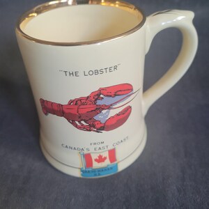 Puede incluir: Una taza de cerámica blanca con un borde dorado y una ilustración de langosta roja. La taza dice "The Lobster" y "From Canada's East Coast".
