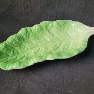 May include: A green ceramic serving dish shaped like a lettuce leaf. The dish is glazed and has a white interior.